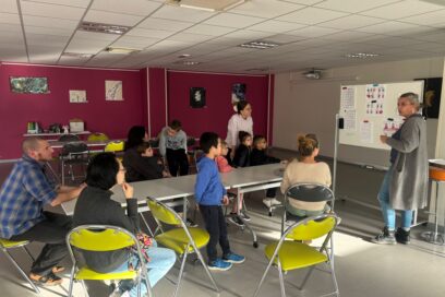 Photo paysage dans une salle, des enfants et parents réunis autour d'une table, une femme à côté d'un tableau blanc désigne des choses dessus - Agrandir l'image, fenêtre modale
