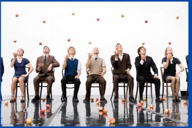 sur une scène aux reflets miroitants argentés, une rangée de chaises face au public, sur lesquelles sont assis 5 circassiens en costumes années 40 en train de jongler avec des oranges , oranges en l'air, oranges éparpillées au sol , formant un charmant tableau joyeux et décalé