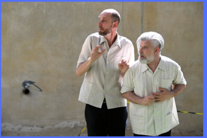 Deux comédiens en manches de chemises blanches se tiennent de bout de profil dans une rue éclairée par la lumière du jour, les mains repliées et jointes dans une mimique identique de surprise et de frayeur leur regard tourné hors champ vers la gauche de l'image - Agrandir l'image, fenêtre modale