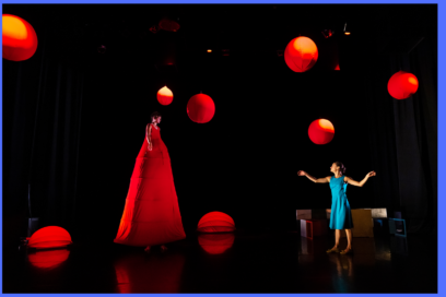 Sur scène plongée dans le noir deux comédiennes en robes de couleur vive, entourée de balles rouges; l'une est géante et dans une robe rouge à gauche, à droite l'autre en robe turquoise près du corps et décolleté, les bras écartés dans un mouvement de danseuse - Agrandir l'image, fenêtre modale