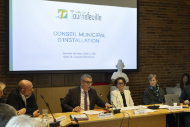 Photo de quatre personnes assises derrière une table, deux hommes et deux femmes, on voit derrière eux le buste de Marianne, et projeté sur une toile "Conseil municipal d'installation"
