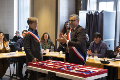 le Maire applaudit debout devant une table sur laquelle il y a une dizaine de boîtes ouvertes avec les écharpes tricolores à l'intérieur. À côté de lui, une élue porte l'écharpe, debout - Agrandir l'image 5 sur 16, fenêtre modale