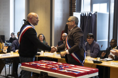 photo du Maire debout qui porte l'écharpe et serre la main à un élu qui porte également l'écharpe - Agrandir l'image 6 sur 16, fenêtre modale