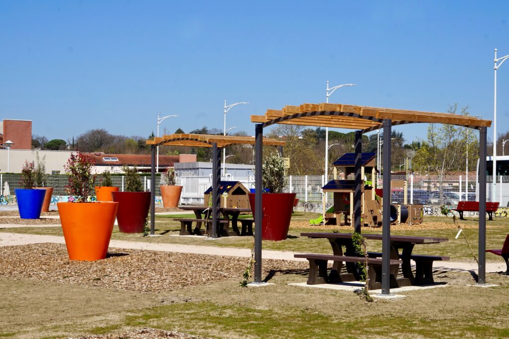 Photo d'un square, grand ciel bleu, avec du mobilier d'extérieur : des grands pots avec des plantes, une table de pique-nique ombragée - Agrandir l'image, fenêtre modale