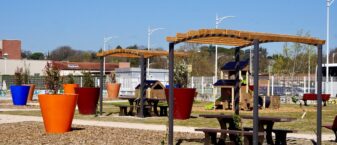 Photo d'un square, grand ciel bleu, avec du mobilier d'extérieur : des grands pots avec des plantes, une table de pique-nique ombragée