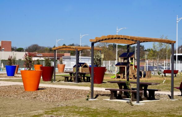 Photo d'un square, grand ciel bleu, avec du mobilier d'extérieur : des grands pots avec des plantes, une table de pique-nique ombragée