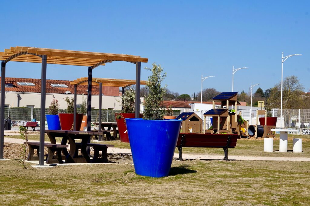 Photo du square éphèmère sous un grand ciel bleu, avec sol végétal, herbe et grandes plantes en pot, zone d'ombre avec pergola - Agrandir l'image, fenêtre modale