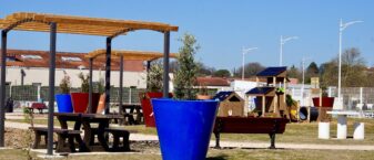 Photo du square éphèmère sous un grand ciel bleu, avec sol végétal, herbe et grandes plantes en pot, zone d'ombre avec pergola