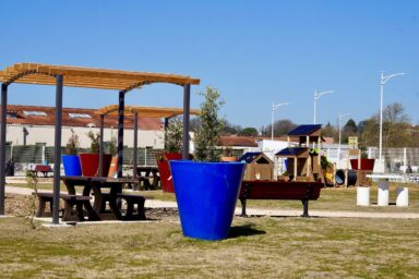 Photo du square éphèmère sous un grand ciel bleu, avec sol végétal, herbe et grandes plantes en pot, zone d'ombre avec pergola