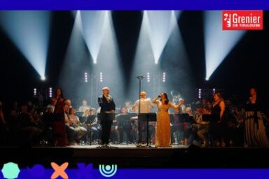 sur scène sous la lumière des projecteurs nimbés de fumée, les chanteurs du Co,ncert du Grenier se tiennent debout au micro face public devant le parterre des musiciens de l'orchestre d'Harmonie