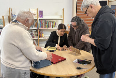 photo dans la Médiathèque, autour d'une table ronde, quatre adultes rassemblés, une femme montre à une autre femme une grille sur la table qui permet d'écrire en braille - Agrandir l'image 3 sur 16, fenêtre modale