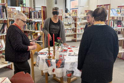 photo à la Médiathèque, autour d'une table préparée comme pour un dîner aux chandelles, trois femmes regardent une quatrième femme qui porte un masque sur les yeux et met la main sur la nappe comme pour chercher quelque chose - Agrandir l'image 4 sur 16, fenêtre modale