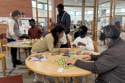 photo à la Médiathèque, deux tables rondes autour desquelles des adultes et des enfants, portant des masques sur les yeux, font un atelier avec des objets répartis sur les tables - Agrandir l'image 2 sur 16, fenêtre modale