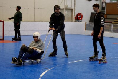 photo dans un gymnase, plusieurs hommes font du roller, un homme pousse un autre assis sur une sorte de chariot - Agrandir l'image 5 sur 16, fenêtre modale
