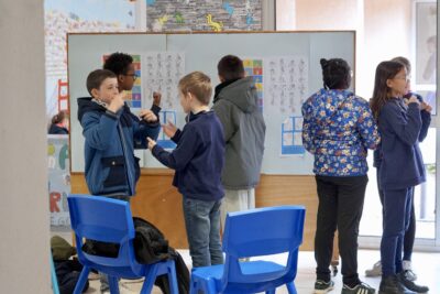 des enfants habillés en bleu font un atelier de langue des signes devant un tableau qui explique les différents signes de main - Agrandir l'image 13 sur 16, fenêtre modale