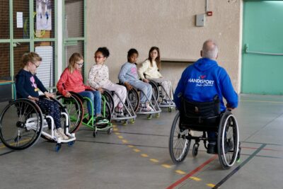 photo de cinq enfants réunis en ligne dans un gymnase, assis dans un fauteuil roulant, avec un homme en fauteuil qui porte un pull avec écrit dans le dos "handisport" - Agrandir l'image 12 sur 16, fenêtre modale