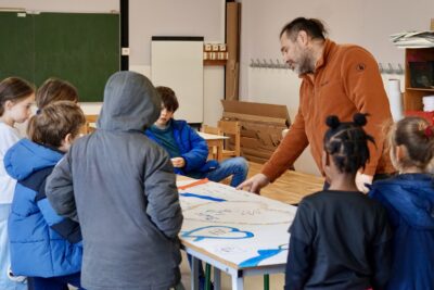 photo d'un atelier avec des jeunes enfants rassemblés autour d'une table, ils sont habillés en bleu, un homme leur désigne quelque chose sur la table, il y a une banderole avec des illustrations de coeur - Agrandir l'image 15 sur 16, fenêtre modale