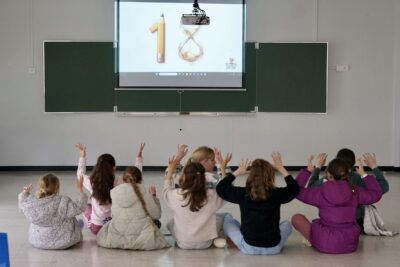 photo de 8 jeunes élèves assis dans une salle de classe, par terre, qui regardent une projection du nombre 18 et lèvent les mains en l'air - Agrandir l'image 16 sur 16, fenêtre modale