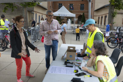 photo de la bourse aux vélos à l'entrée, avec deux bénévoles derrière une table qui saluent le Maire et une élue, sur une place en extérieur - Agrandir l'image 1 sur 19, fenêtre modale