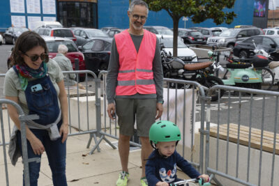 photo d'un petit enfant qui teste un vélo à roulettes, avec une femme derrière lui et un homme qui porte un gilet rose - Agrandir l'image 11 sur 19, fenêtre modale