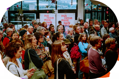 Foule rassemblée de profil dans un hall lumineux pendant les festival Marionnettissimo, que signale un affichage sur les baies vitrées en arrière-plan de la photo. - Agrandir l'image, fenêtre modale