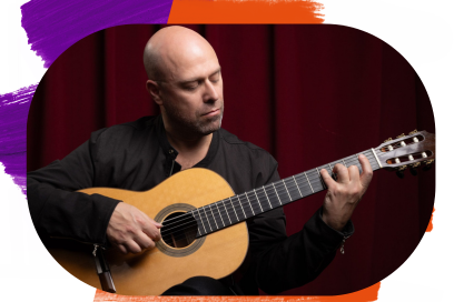 L’image montre un homme chauve assis en train de jouer d’une guitare classique acoustique. Il porte une veste noire et semble concentré, les yeux fermés ou baissés vers l’instrument. La guitare a une finition en bois clair avec un manche sombre, et sa main gauche forme un accord assez haut sur le manche pendant que sa main droite pince les cordes près de la rosace. L’arrière-plan est composé de rideaux rouges foncés. - Agrandir l'image, fenêtre modale