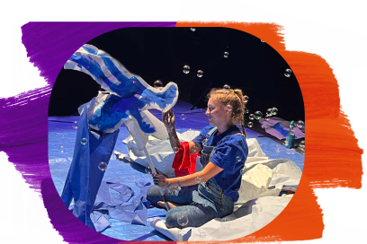 Sur scène, cernée de bulles de savon voletant dans l'air une comédienne souriante en manches de chemise bleue se tient de profil agenouillée manipulant une baudruche en plastique bleu représentant un dragon. - Agrandir l'image, fenêtre modale