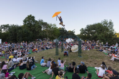 photo d'un spectacle en plein air dans un parc, au centre un homme suspendu à une sorte de parapluie, plusieurs mètres au-dessus du sol, tout autour, de nombreux spectateurs, familles notamment, qui regardent