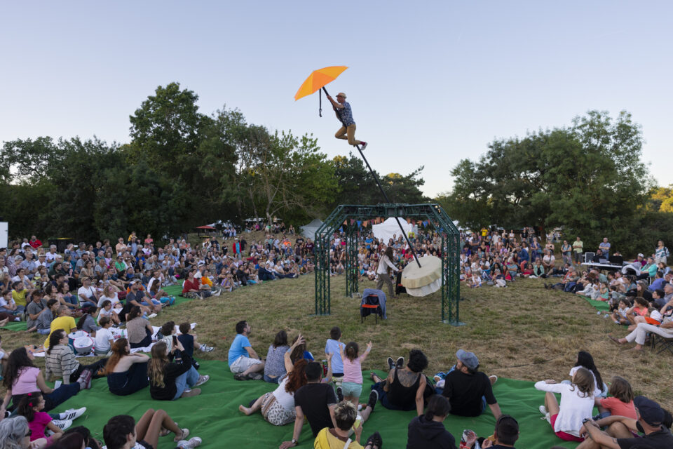 photo d'un spectacle en plein air dans un parc, au centre un homme suspendu à une sorte de parapluie, plusieurs mètres au-dessus du sol, tout autour, de nombreux spectateurs, familles notamment, qui regardent