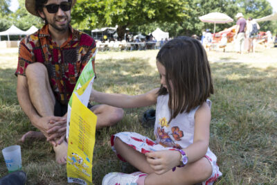 photo d'une petite fille assise dans l'herbe à côté d'un homme, elle regarde le dépliant des excentriques - Agrandir l'image 9 sur 15, fenêtre modale