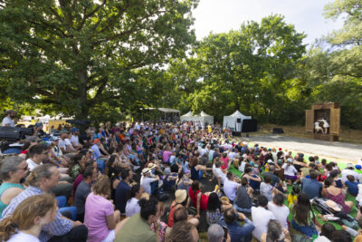 photo d'un spectacle vu depuis l'arrière du public, nombreux, assis à l'ombre dans le parc pour assister au spectacle. Sur scène, deux personnes habillées en blanc dans une grande armoire ouverte - Agrandir l'image 13 sur 15, fenêtre modale