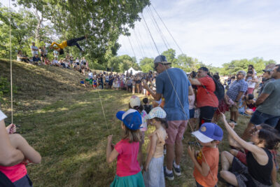 photo d'un spectacle d'acrobatie, une personne est suspendue dans les airs par des fils tenus par le public, dont de nombreux enfants, le tout dans un parc au soleil - Agrandir l'image 11 sur 15, fenêtre modale