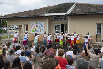photo d'un spectacle devant la maison de quartier, une vingtaine de jeunes femmes dansent en tenue tee shirt blanc et pareo/robe colorée, couronne de fleurs sur la tête, les bras écartés et alignées en trois lignes sur scène devant les spectateurs nombreux, assis devant la scène - Agrandir l'image 8 sur 14, fenêtre modale