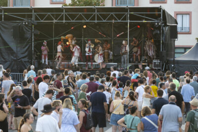 photo de la scène vue du public, avec un public familial nombreux, et un groupe avec une dizaine de musiciens en costumes blancs et rouges - Agrandir l'image 3 sur 14, fenêtre modale