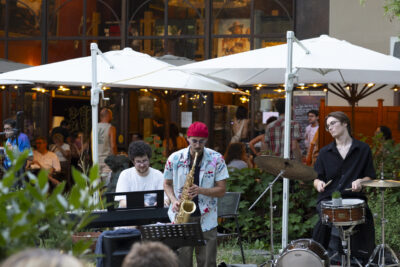 photo d'un concert en extérieur, un groupe de trois personnes avec un au clavier, un à la batterie et un au saxophone, et le public au premier plan de dos - Agrandir l'image 4 sur 14, fenêtre modale