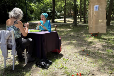 photo de deux femmes en face à face assises à une table au soleil, dans un parc, une porte une tenue de voyante - Agrandir l'image 16 sur 19, fenêtre modale
