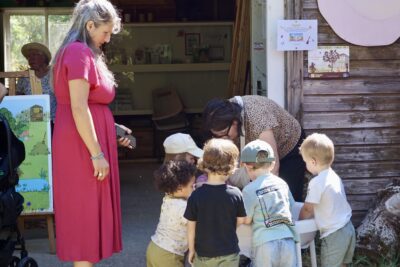 photo de quatre petits enfants de dos, en extérieur, avec une femme à côté qui les regarde faire quelque chose sur une table cachée - Agrandir l'image 8 sur 19, fenêtre modale