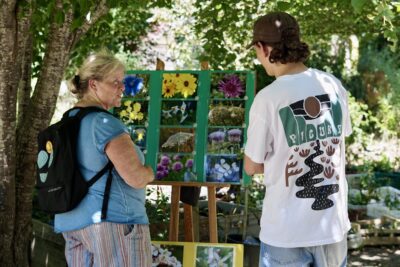photo d'une femme et d'un jeune homme de dos, qui observent un panneau avec des photos de fleurs accrochées, en extérieur à l'ombre - Agrandir l'image 15 sur 19, fenêtre modale