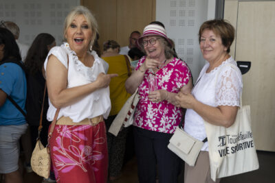 photo de trois femmes, une à droite montre du doigt celle sur sa gauche, qui montre du doigt l'objectif. Toutes trois sourient et ont l'air de bonne humeur, dans une salle, on voit d'autres personnes de dos derrières elles - Agrandir l'image 13 sur 13, fenêtre modale