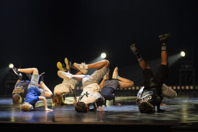 Sur la scène du phare, en tee shirts amples et colorés et bermudas, les danseurs de hip hop effectuent une figure au sol, mains en appui et jambes dressées en l'air. - Agrandir l'image 2 sur 9, fenêtre modale