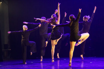 Sur la scène nimbée de lumière violette des danseuses en robe noire à volants effectuent une chorégraphie collective, bras levés au ciel, en appui sur la pointe du pied gauche, de dos à la caméra. - Agrandir l'image 3 sur 9, fenêtre modale