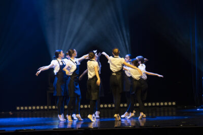 Un groupe de danseuses aux chignons serrés, en tunique noire à bustier et dos nu sont rassemblées en cercle au centre de la scène, des rais de lumière bleus pulsent en fond de scène un rideau qui semble émaner de ce cercle. - Agrandir l'image 4 sur 9, fenêtre modale