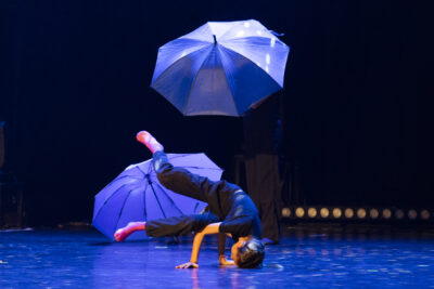 Sur scène, dans une atmosphère bleutée qui évoque un temps de pluie, une danseuse de hip hop effectue une figure au sol, en appui sur les mains et les pieds jetés en arrière en tenue noire sobre, des parapluies ouverts encadrent la scène. - Agrandir l'image 7 sur 9, fenêtre modale