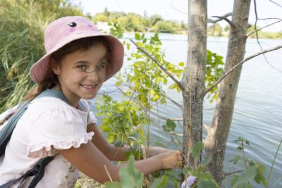 photo en extérieur d'une enfant qui sourit à l'objectif, en extérieur à côté d'un lac, elle prend quelque chose d'un arbre - Agrandir l'image 17 sur 21, fenêtre modale