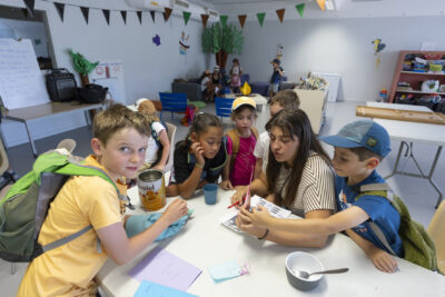 photo d'un groupe d'une dizaine d'enfants autour d'une table, avec des sacs à dos, en train de regarder une feuille avec une animatrice - Agrandir l'image 18 sur 21, fenêtre modale