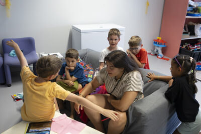 photo dans une salle de 5 enfants avec une animatrice, qui s'amusent autour d'un canapé gris - Agrandir l'image 21 sur 21, fenêtre modale
