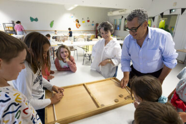 photo d'un groupe de plusieurs enfants autour d'une table sur laquelle est posée un jeu en bois, dans une grande pièce, une jeune fille joue avec le Maire