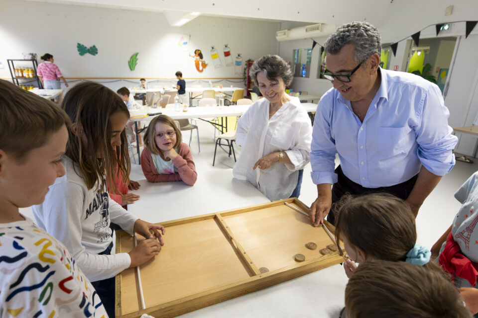 photo d'un groupe de plusieurs enfants autour d'une table sur laquelle est posée un jeu en bois, dans une grande pièce, une jeune fille joue avec le Maire