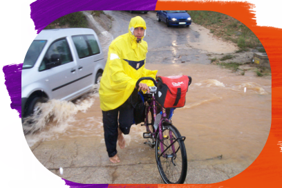 Sur une route inondée d'eau boueuse, un homme en toile cirée jaune, se tient face caméra tenant à la main une bicyclette aux bagage rouge en toile cirée également, derrière lui circulent des voitures faisant jaillir des éclaboussures de sous leurs pneus - Agrandir l'image, fenêtre modale