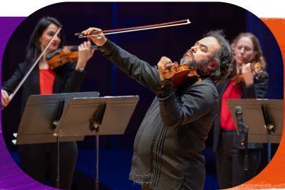 Photographie de concert montrant le violoniste Alexis Cárdenas au premier plan, de profil, levant son archet bien au-dessus de son violon dans un geste expressif. Il porte une chemise noire plissée. En arrière-plan, deux violonistes en veste noire et haut rouge l'accompagnent derrière des pupitres. L'ambiance lumineuse est sombre et feutrée. - Agrandir l'image, fenêtre modale